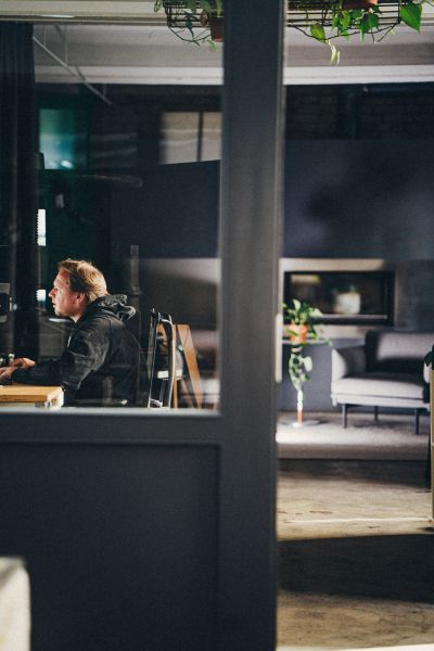 Eine Person im Hoodie sitzt am Schreibtisch in einer Agentur, durch eine Tür-Glasscheibe fotografiert